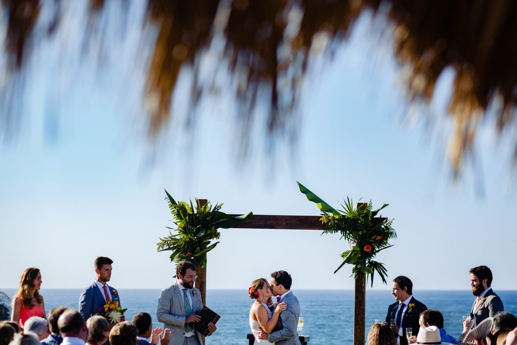 bride and groom kissing during the ceremony  with the ocean on the background and the guest looking at them 