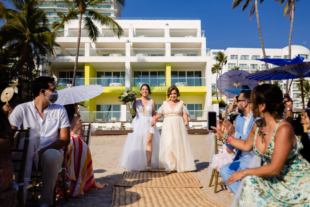 bride and her mum walk to the ceremony with the Hilton hotel on the background her with the bouquet on her hand all the people are looking them