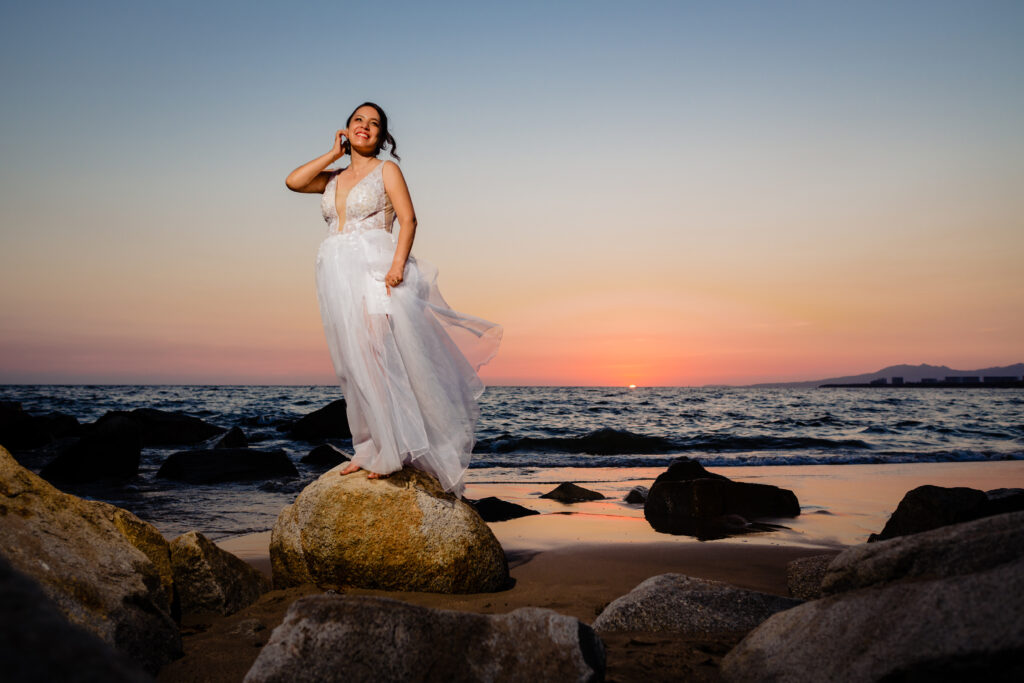 bride on the beach posing on a rock with ocean and sunset on the background