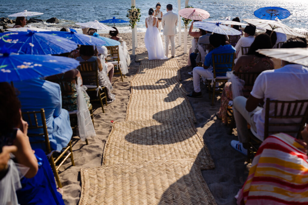 bride and groom on the ceremony on the beach with the ocean on the background and all the guest with umbrellas 