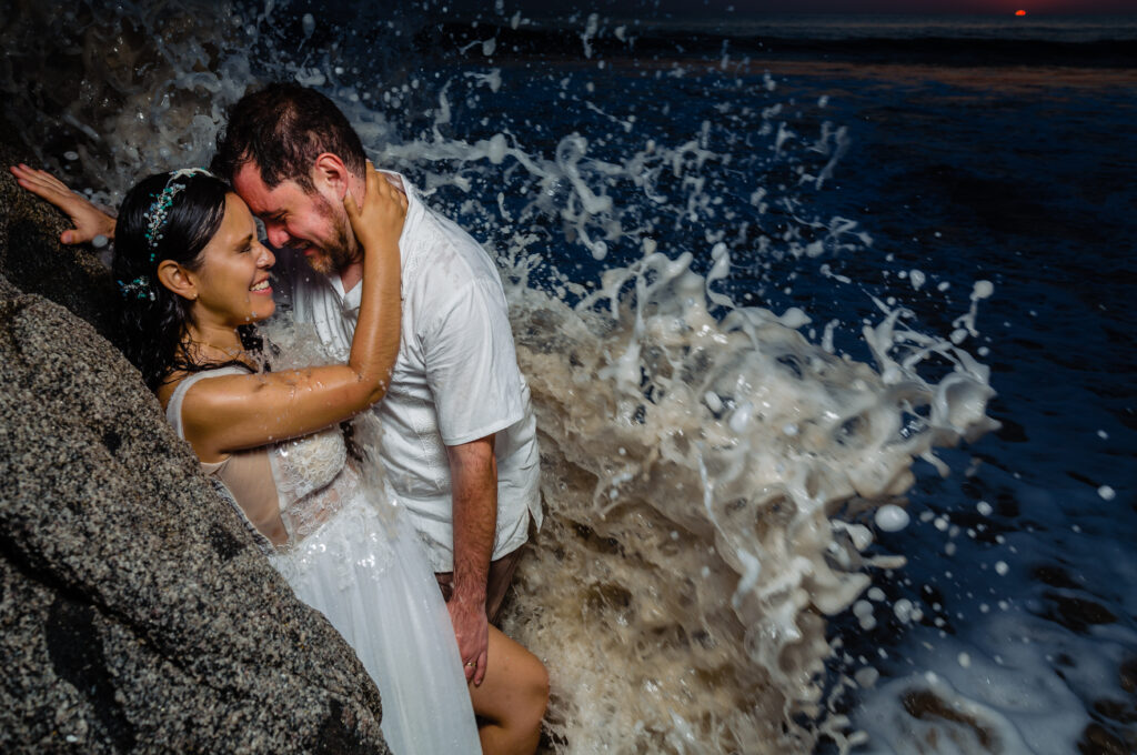 bride and groom hugs each other on a rock with a splash of a wave who wet them with the ocean and the sunset on the background 