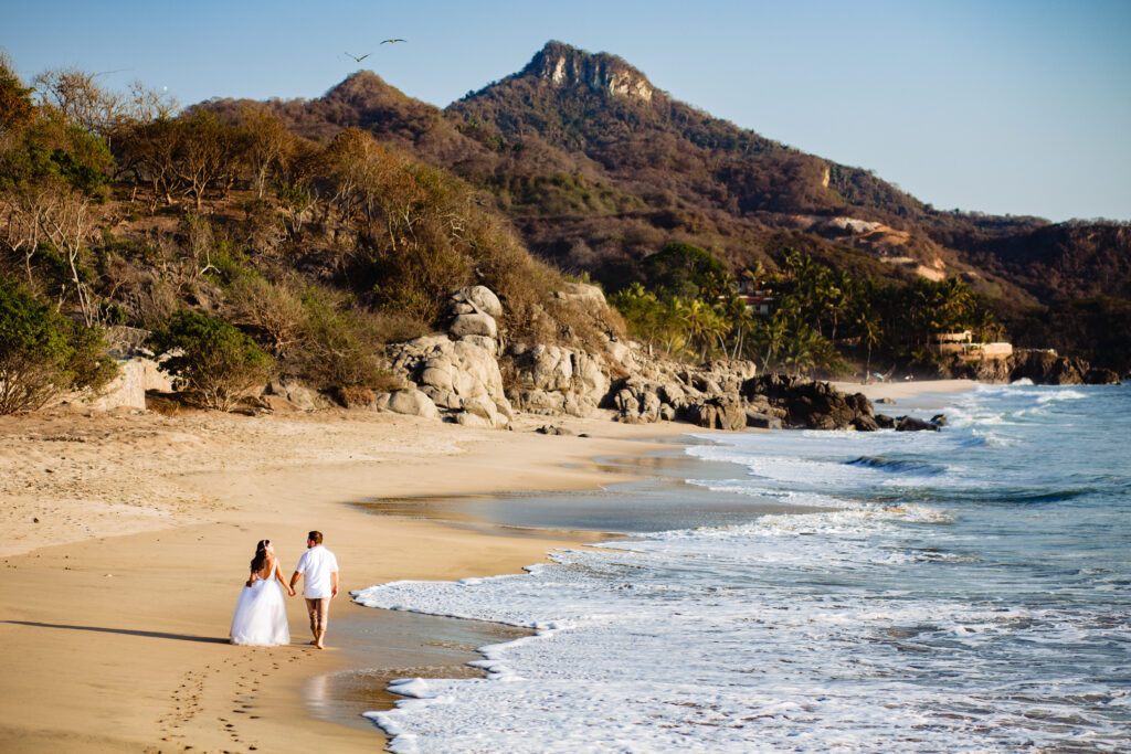 bride and groom walking on the beach holding hands with mountain on the background